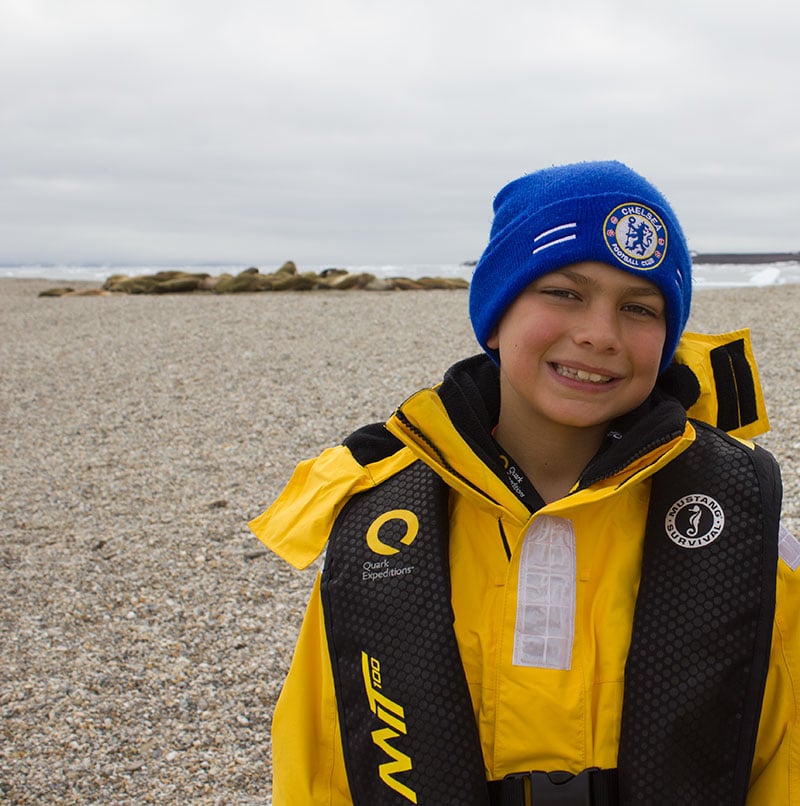 Owen Schuster smiles wide for the camera, a large gathering of walrus hauled out on the Spitsbergen beach behind him. 